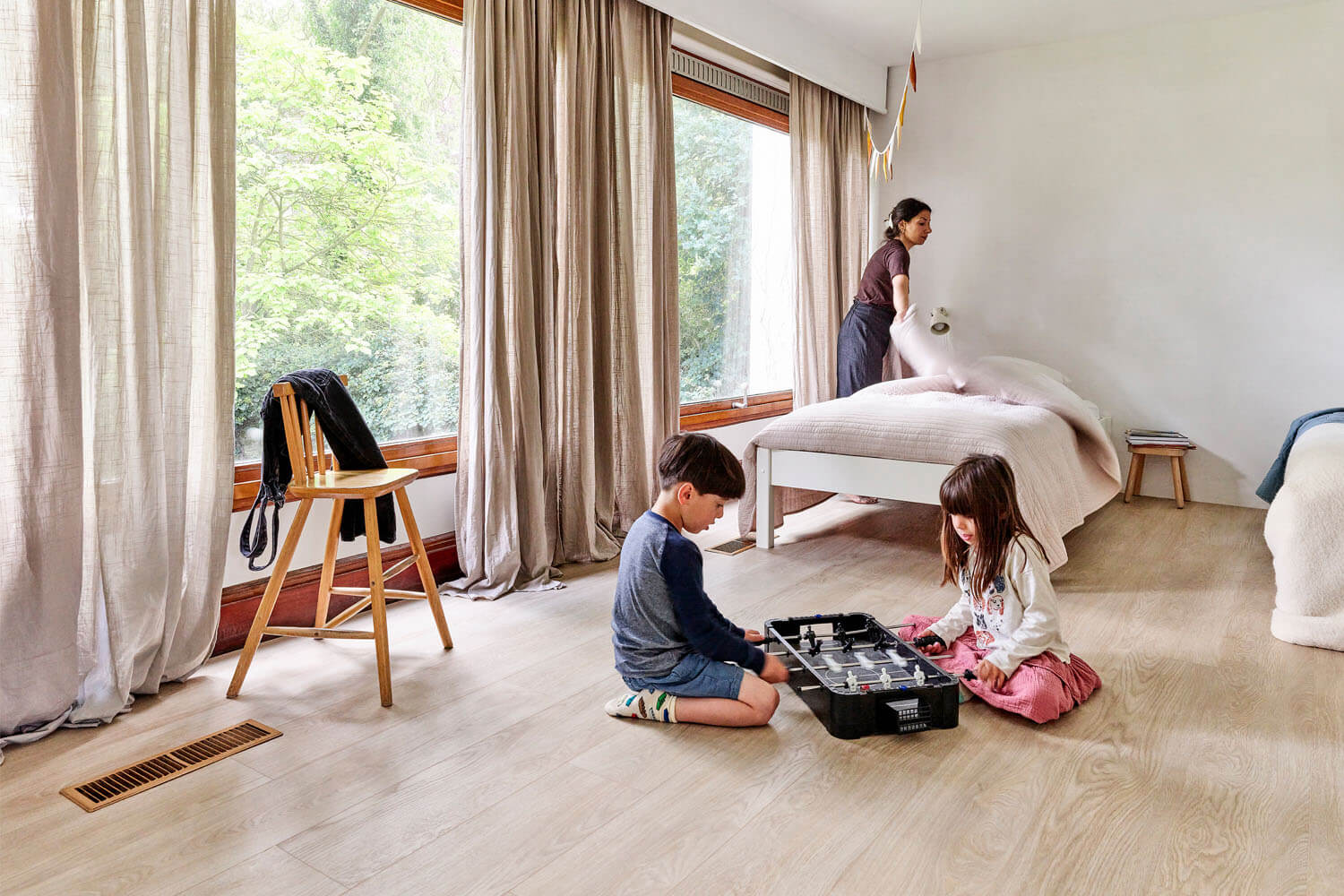Sumeyya maakt het bed op in de slaapkamer van de kinderen, terwijl de kinderen een spelletje spelen op de vloer. Deze vloer is de Moduleo LayRed Laurel Oak 51230 Embossed luxe pvc vloer.
